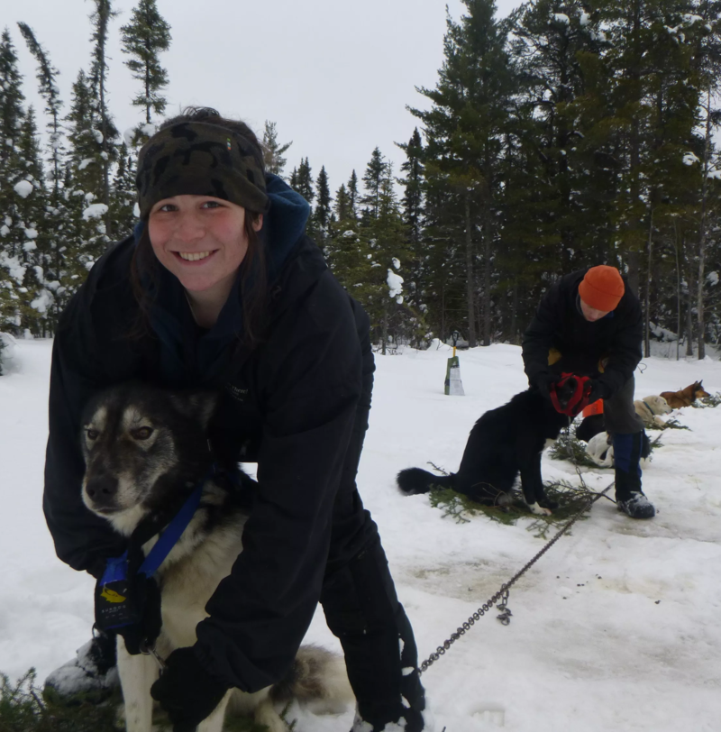 A woman in a black jacket and camouflage beanie smiles while hugging a husky dog with a blue collar. They are in a snowy, wooded area. In the background, another person in an orange hat is tending to other dogs that are leashed to a line. The scene suggests a dog sledding or winter activity setting.